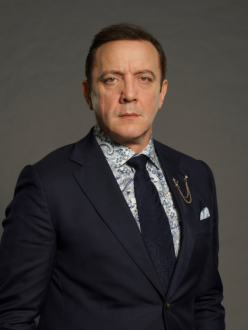 Man in a dark suit, patterned shirt, and tie, with a serious expression, stands against a plain grey background in studio lighting.