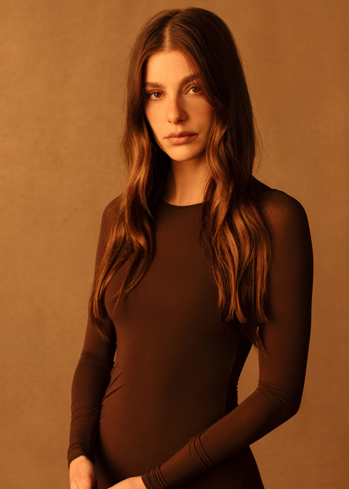 Camila Morrone wears a brown dress in front of a gold backdrop.