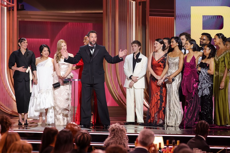 A group of people in formal attire stand on stage at an awards ceremony, with one man speaking at a microphone and others gathered around him, surrounded by a decorated, well-lit event venue with an audience visible in the foreground.