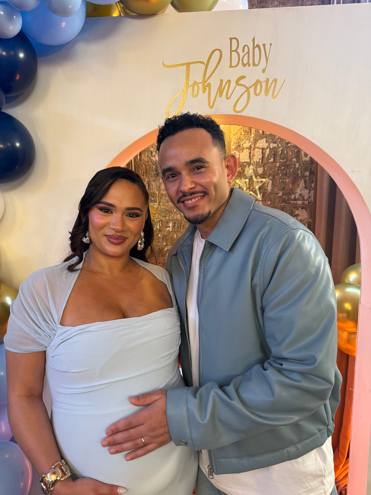 A smiling couple poses at a baby shower in front of a decorated backdrop with "Baby Johnson" written on it, surrounded by balloons. The man gently touches the woman's pregnant belly, creating a joyful, celebratory mood.