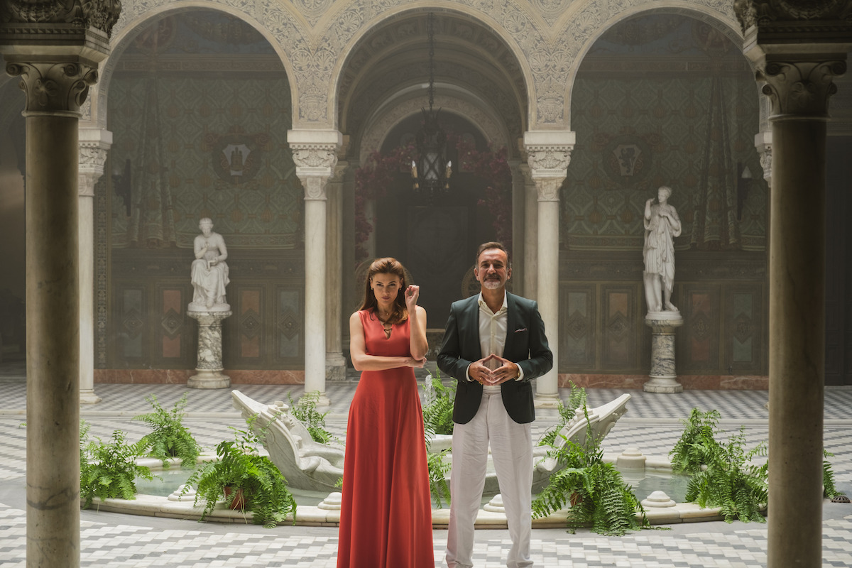 A woman in a red dress and a man in a suit stand in an ornate courtyard with marble columns, statues, a patterned floor, plants, and arched ceilings, creating a formal and elegant setting.