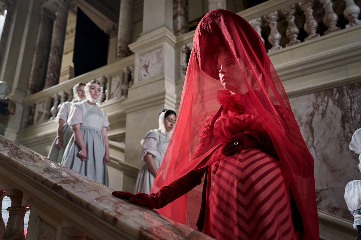 A woman in a dramatic red dress and veil stands on marble stairs in an ornate building, with three women in white dresses behind her. The mood is mysterious and formal, with rich colors and historic architecture.