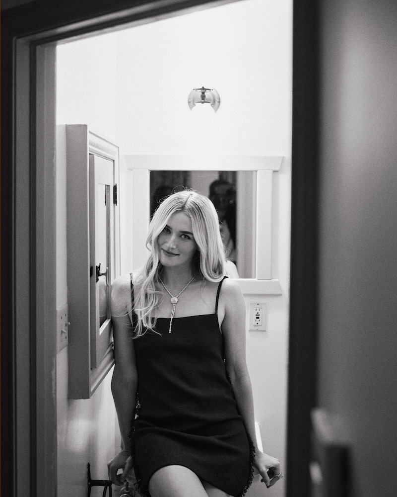 A woman with long blond hair in a black dress sits on a counter in a small, bright bathroom. The scene is framed by a door, with a mirror and wall fixtures visible in the background. Black-and-white photograph.