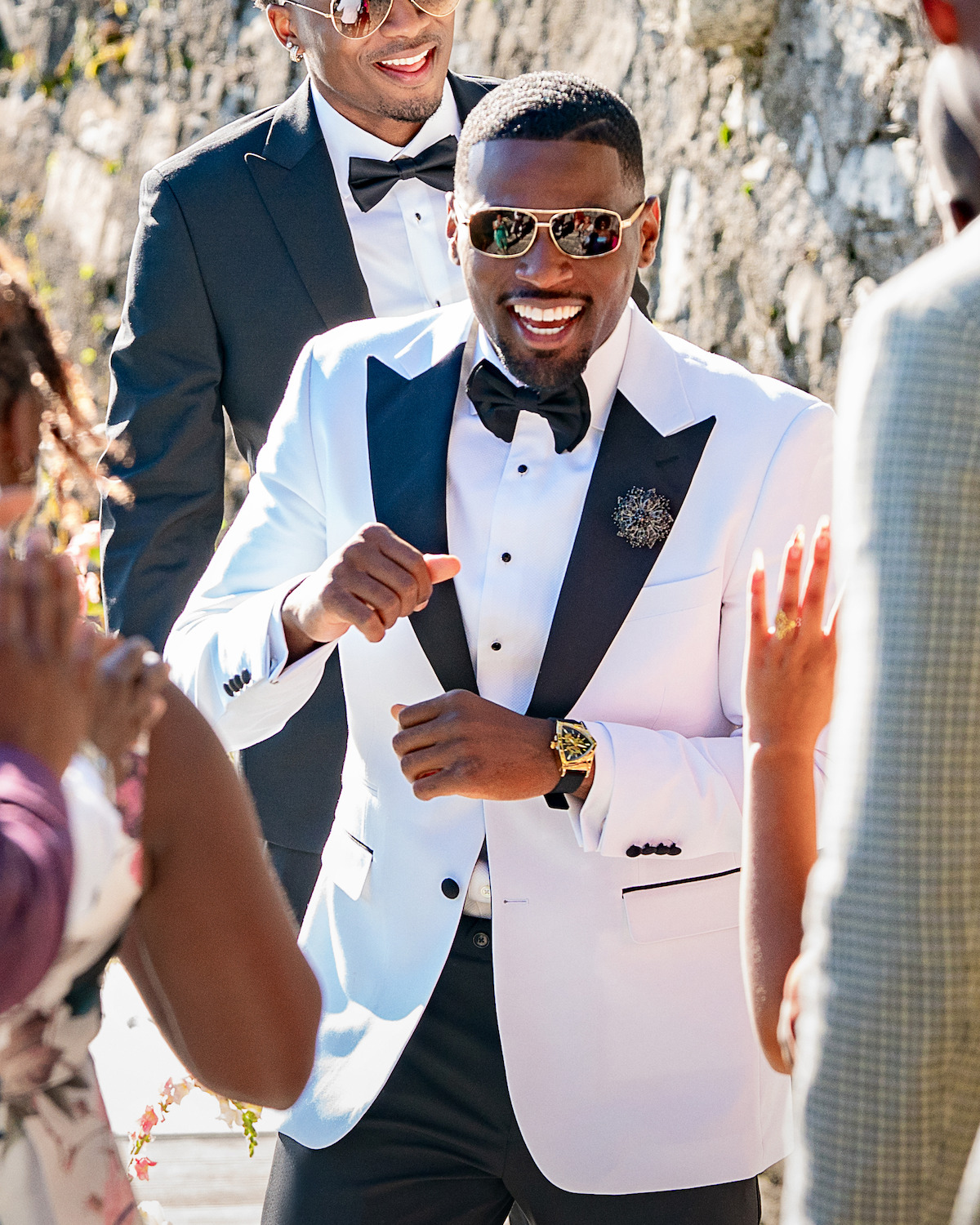 Smiling man in a white tuxedo and sunglasses dancing at an outdoor celebration, surrounded by people in formal attire, with sunlight and a stone wall in the background.