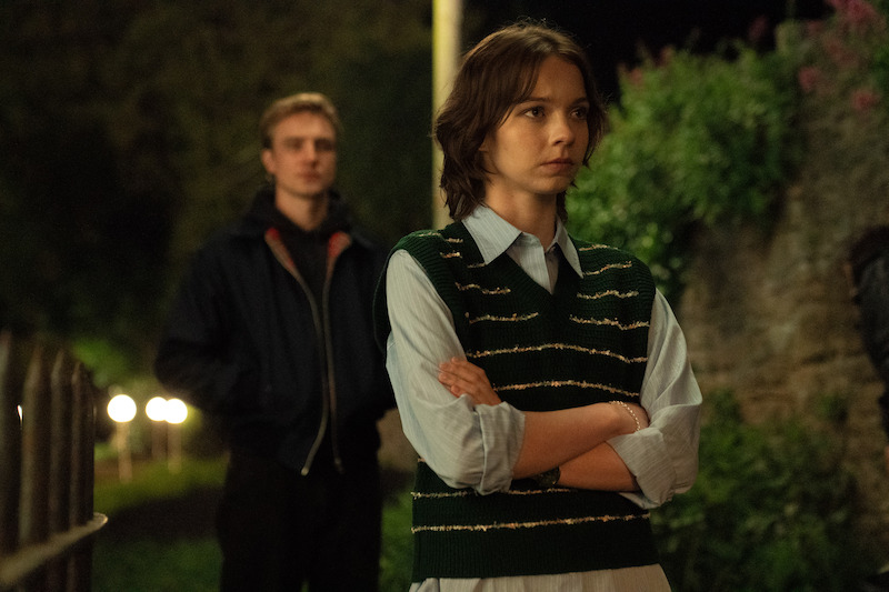 Two young people standing outside at night, one in the foreground with arms crossed looking thoughtful, the other slightly blurred in the background. The environment is dimly lit with greenery and a stone wall visible.