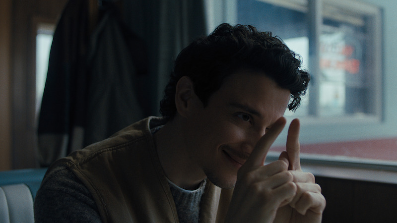 Man with dark curly hair sitting in a diner booth, smiling while holding up both hands with index fingers raised, seen against a window and coat in the background, in natural light.