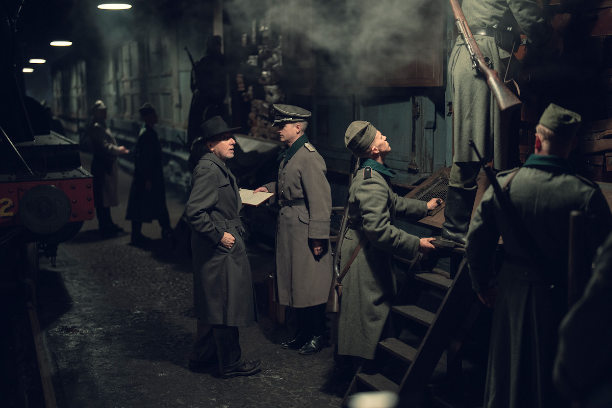 Soldiers in historical uniforms and a man in a hat stand beside a train in a dimly lit, smoke-filled railway station, checking papers and inspecting the train as others climb stairs into a boxcar.