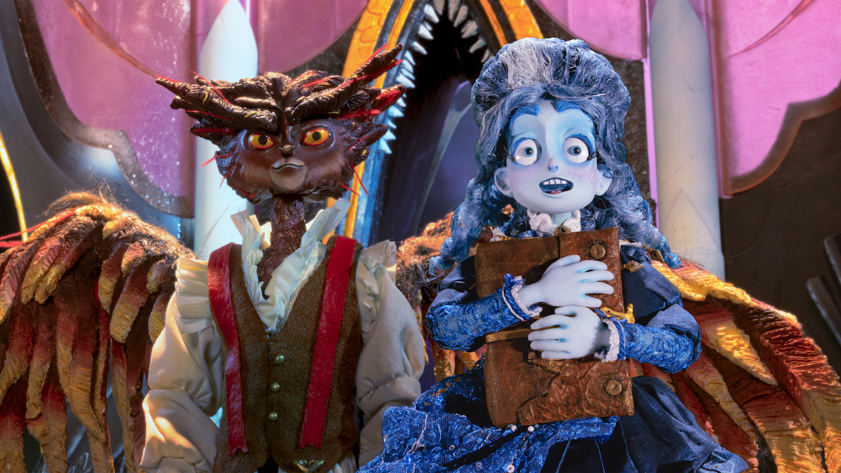 Two detailed stop-motion puppets in fantasy costumes stand in a colorful, ornate environment. One has owl-like features and wings, the other has blue hair and holds a large book, both in front of an elaborate, arched set piece.