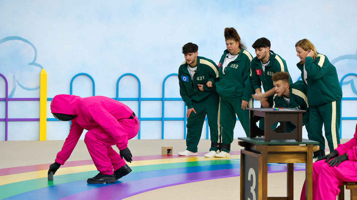 People in green tracksuits watch a masked person in a pink suit kneeling on a colorful, rainbow-striped floor. The scene is bright, playful, and tense, resembling a game show with a whimsical yet competitive atmosphere.