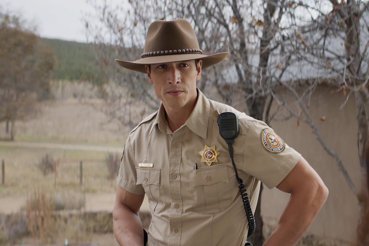 A law enforcement officer in a tan uniform and cowboy hat stands outdoors near a rural building with trees and open land in the background, looking directly at the camera.