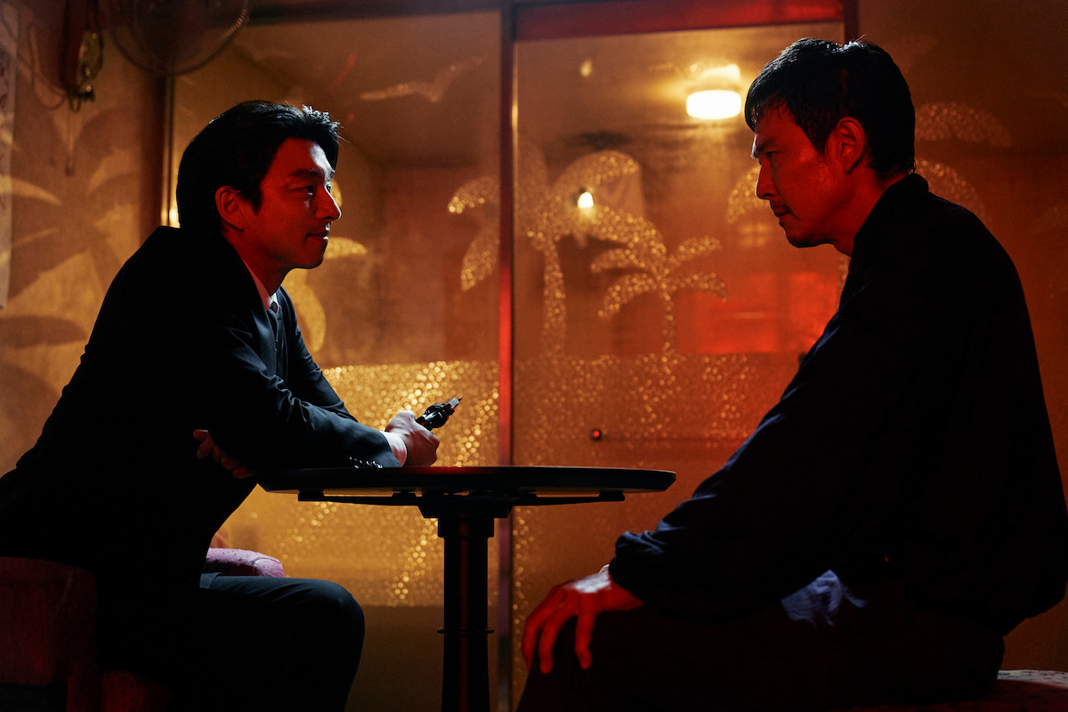 Gi-hun (Lee Jung-jae) sits at a table wearing a grim expression across from another character in a red-lit room.
