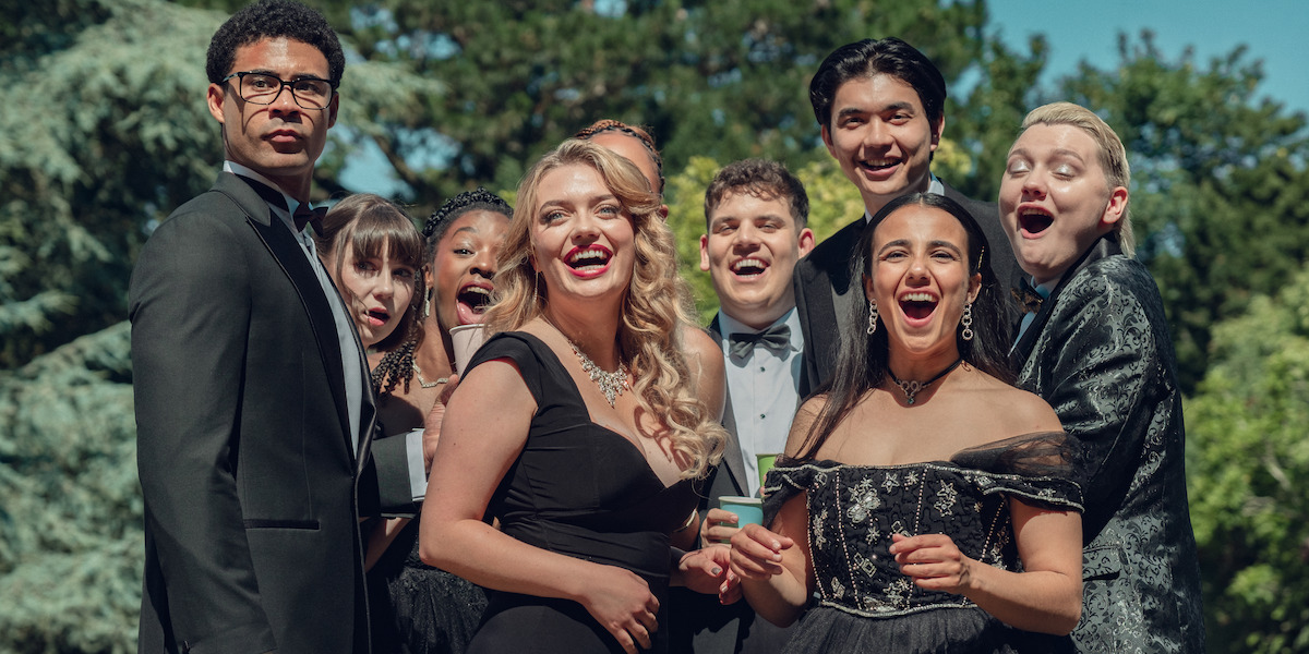 Group of young adults in formal attire posing outdoors, smiling and laughing together for a photo, with green trees and blue sky in the background, suggesting a celebratory event like prom or a formal party.