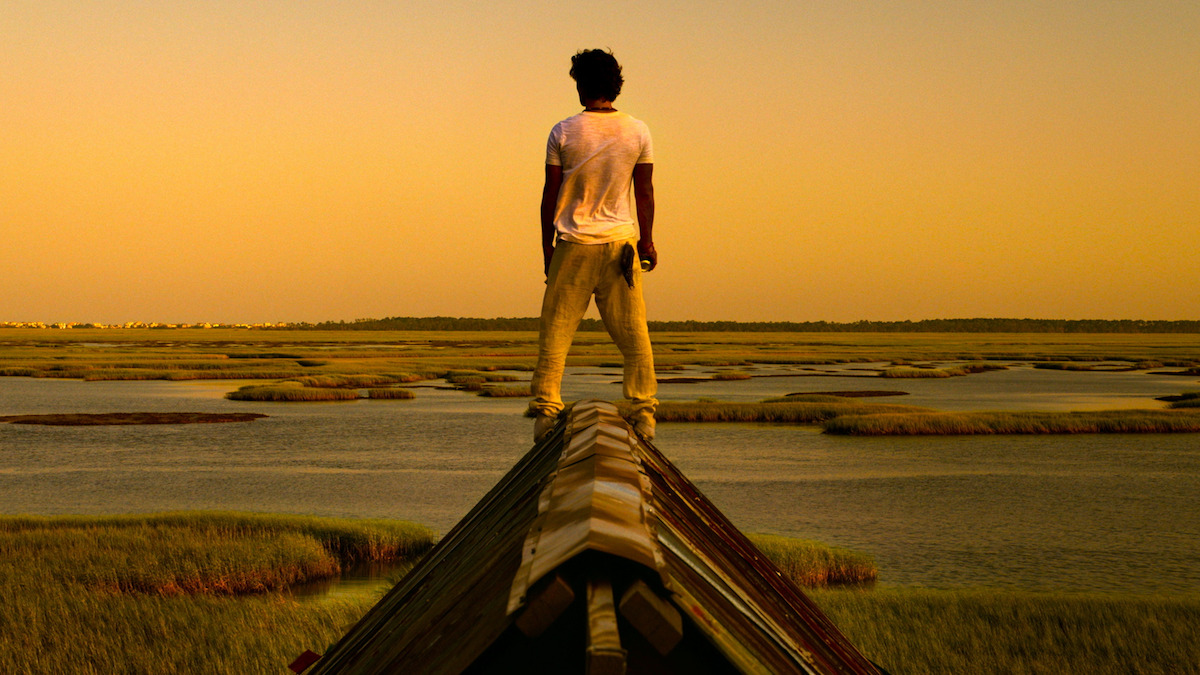 A person stands on a rooftop overlooking a marshland during sunset, surrounded by water and grassy patches under an orange sky.