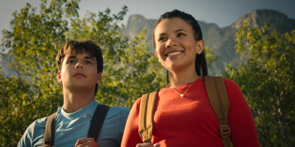 Two young hikers with backpacks stand outdoors, smiling and looking ahead, surrounded by green trees with mountains and blue sky in the background, suggesting adventure in nature.