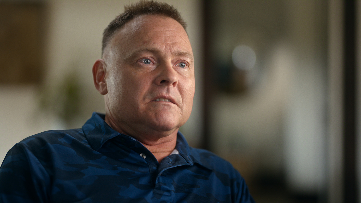 Man with short hair in a blue shirt sits indoors, looking slightly off camera with a serious expression; background is softly blurred, suggesting a home or office environment.