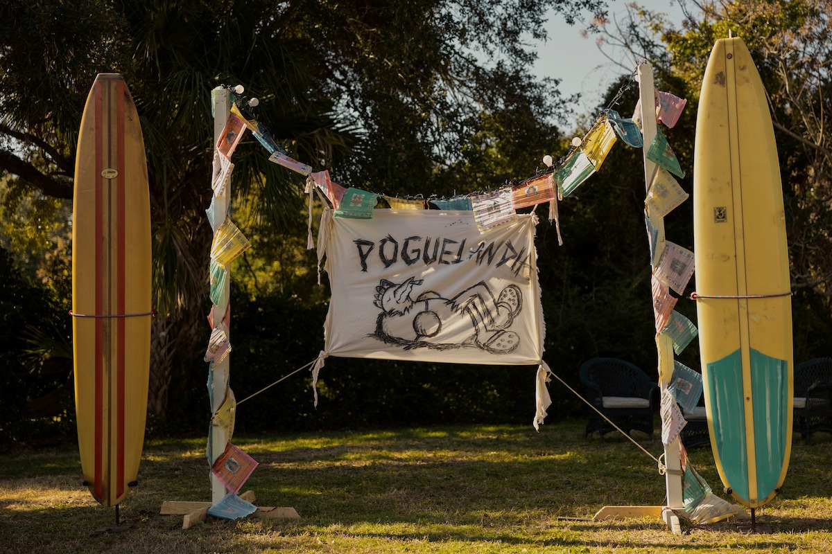 Two colorful surfboards stand upright outdoors with festive garlands and a hand-drawn “POGUELANDIA” banner between them, surrounded by trees and outdoor chairs on grass in a sunny backyard setting.