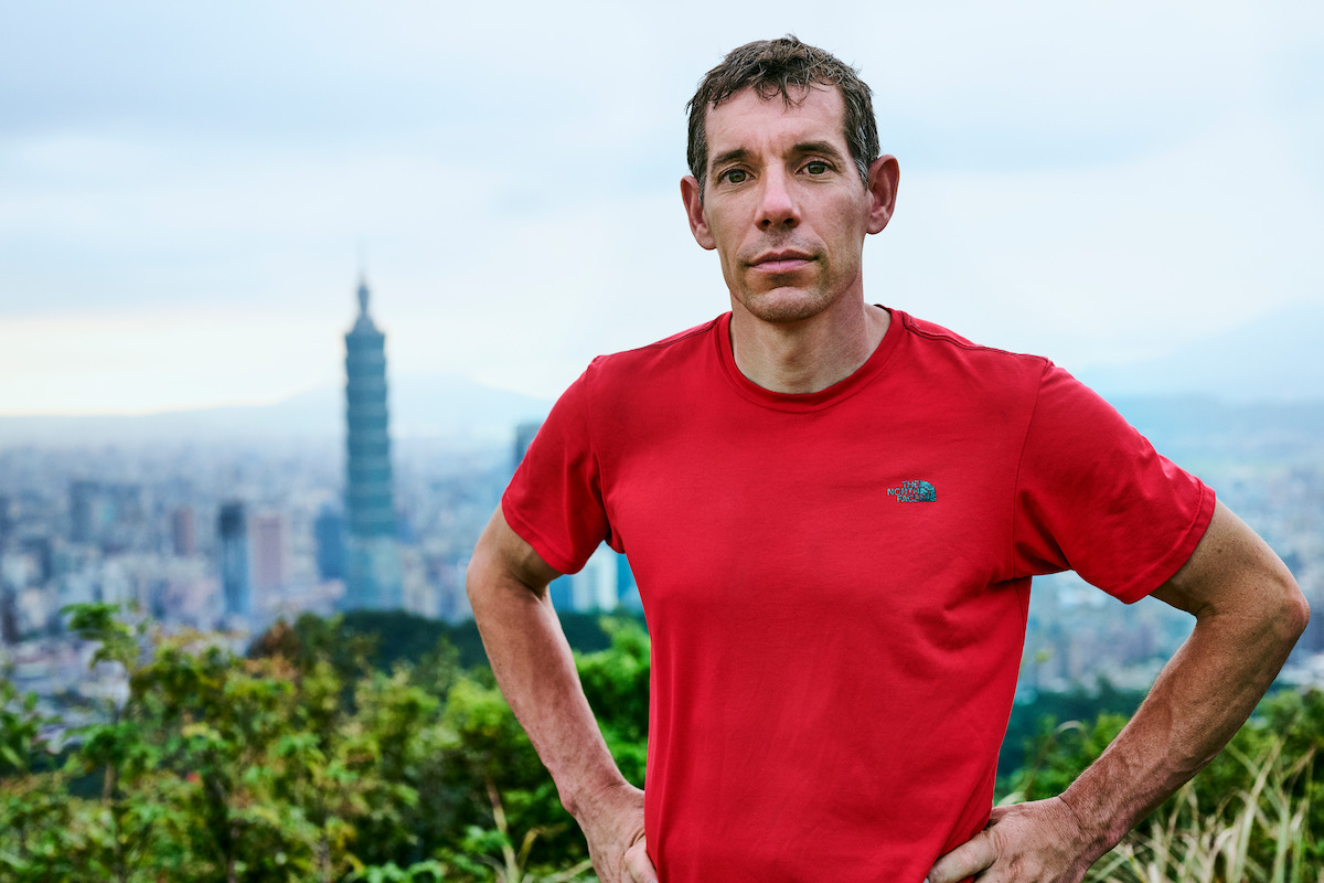 Man in a red shirt stands confidently outdoors with greenery and a cityscape featuring Taipei 101 in the background, under an overcast sky, creating a focused and contemplative mood.