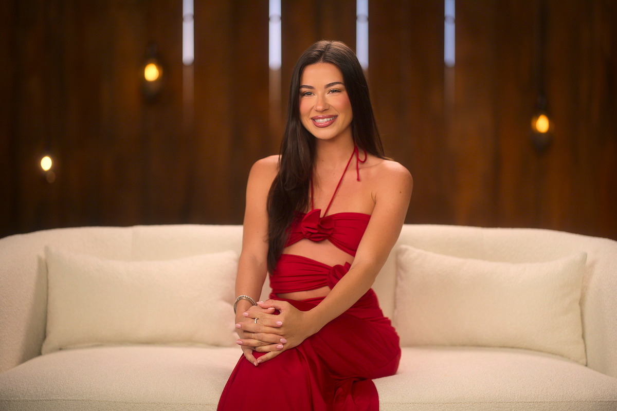 Woman in a red dress sitting on a white couch, smiling, with wooden wall and warm hanging lights in the background, creating a cozy, elegant indoor atmosphere.