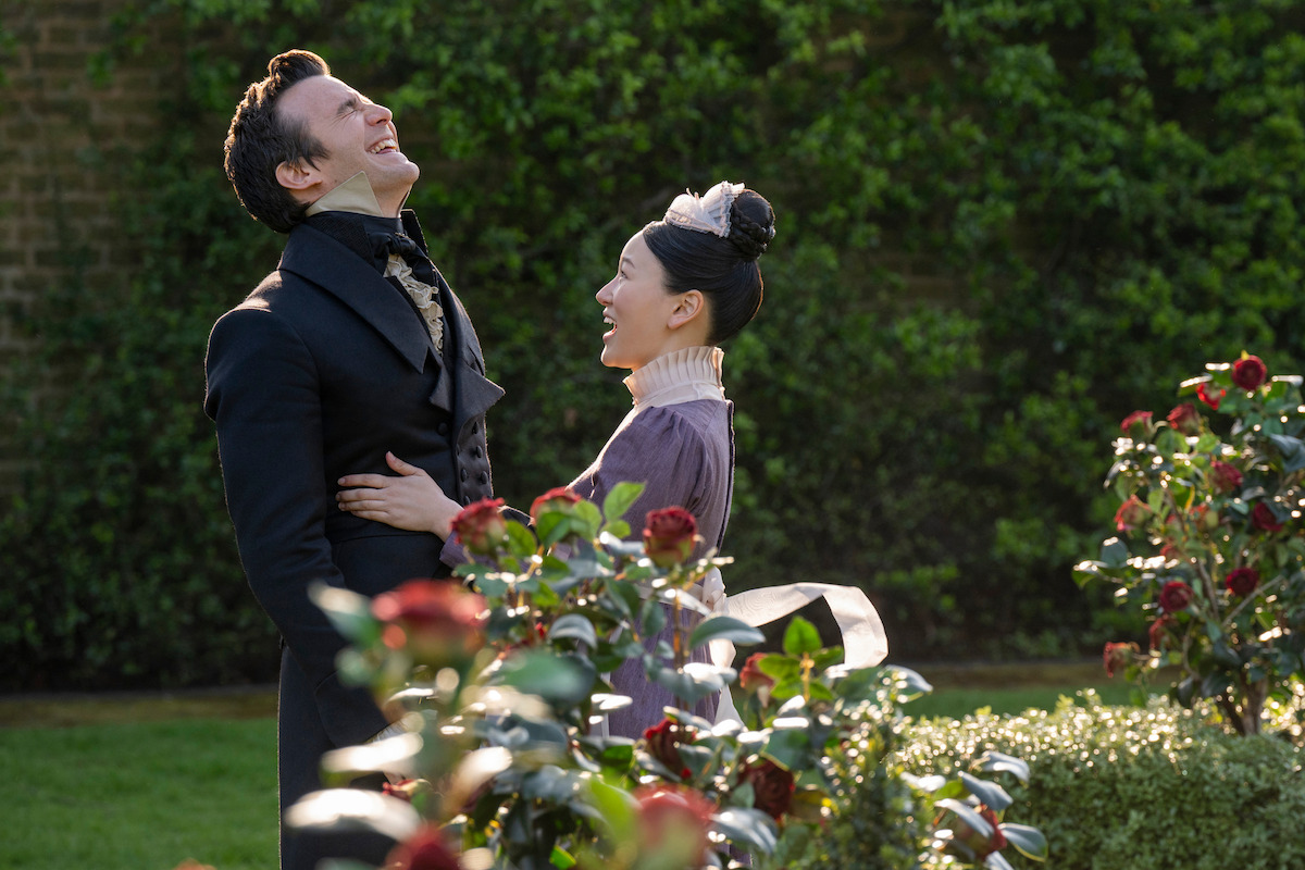 A man and woman in period costumes laugh together in a formal garden with blooming red roses and manicured greenery, under daylight.