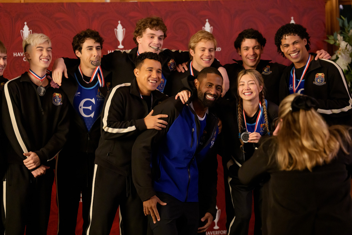 A group of young athletes in matching tracksuits poses happily with medals, smiling for a photo in front of a trophy-patterned red backdrop at an indoor awards ceremony. A woman faces them, appearing to take the picture.