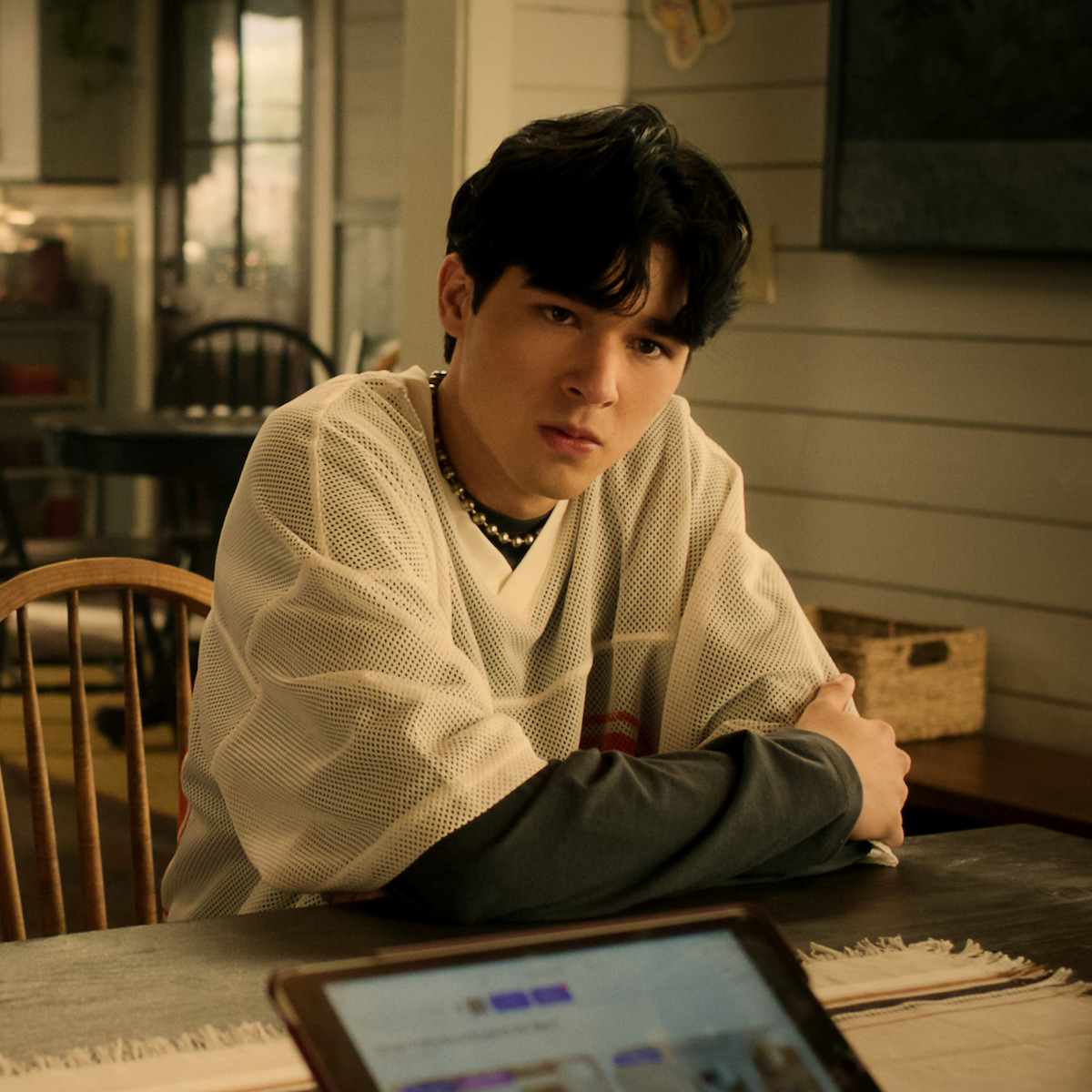 Young person with dark hair sits at a wooden table in a cozy, well-lit room with paneled walls and homey decor, facing a laptop or tablet screen in a casual, modern home environment.
