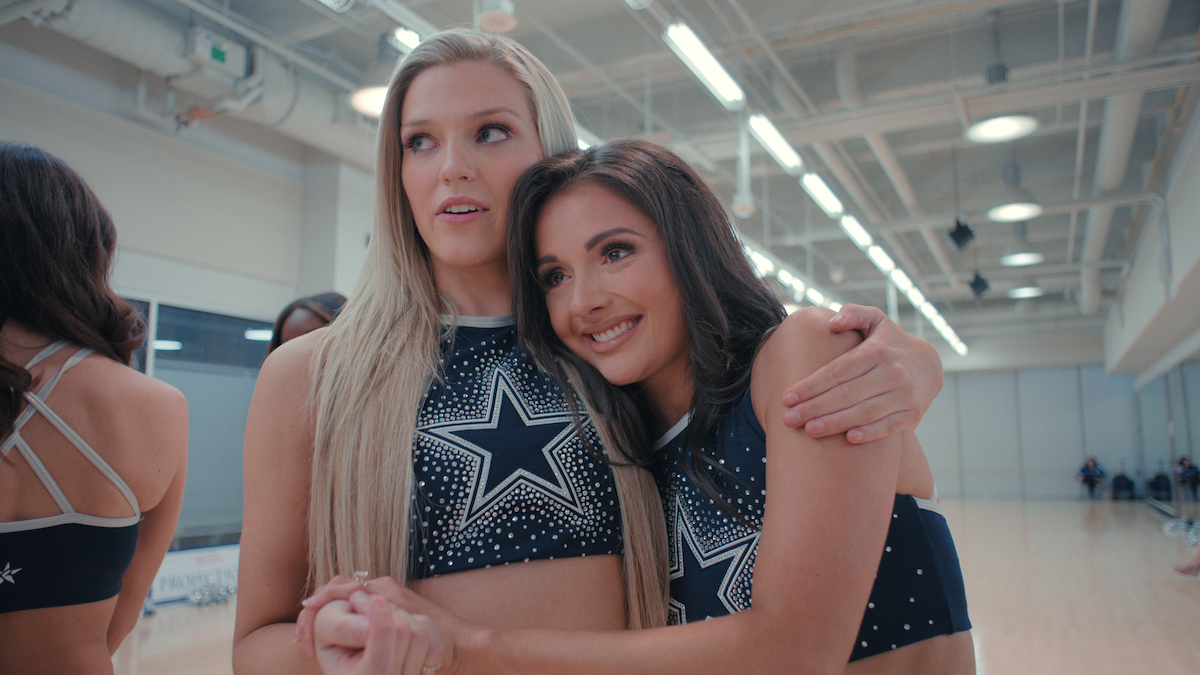 A brunette woman embraces a blonde woman who looks off into the distance, both wearing DCC practice uniforms in an rehearsal space.