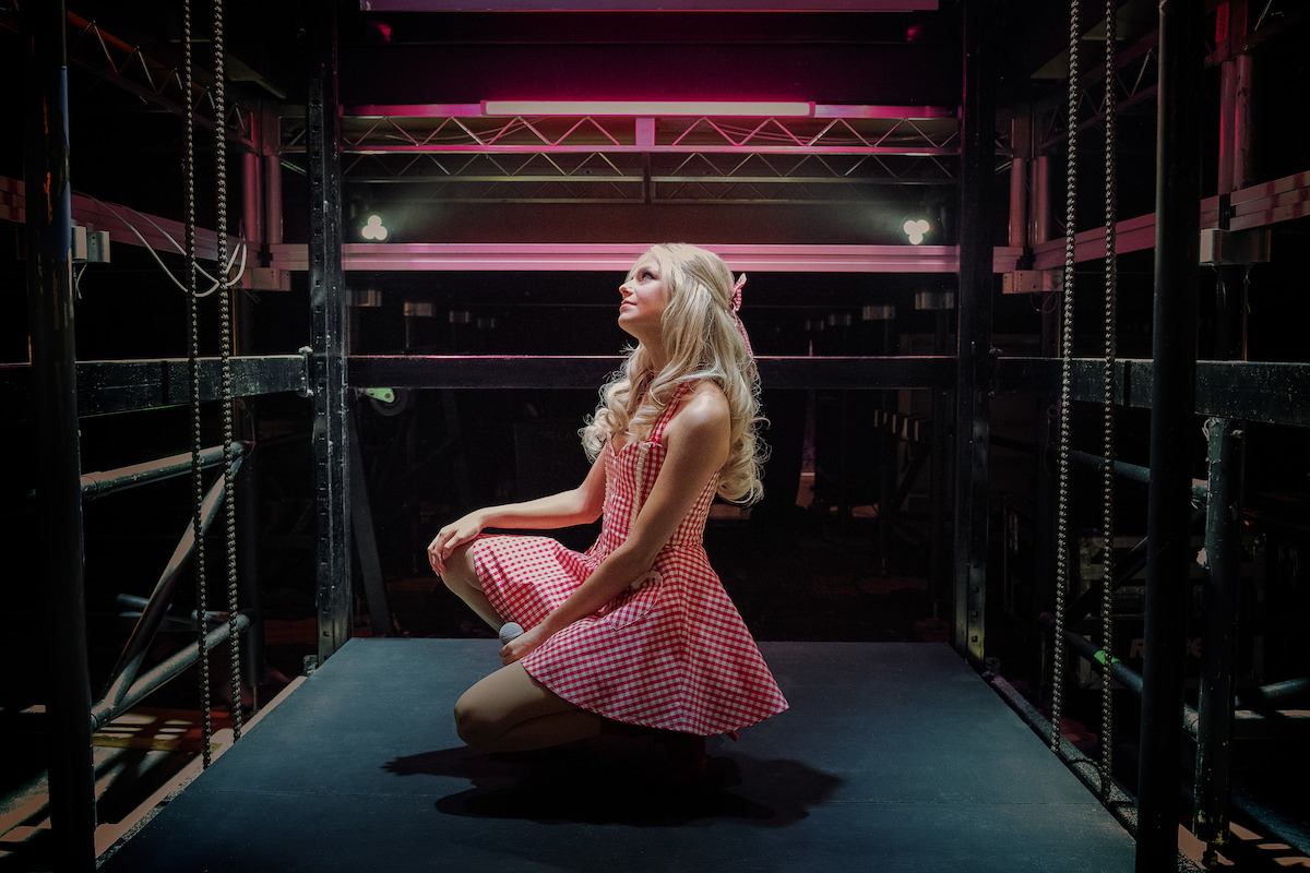Woman in a pink checkered dress kneeling on a stage or backstage area, with dramatic lighting and industrial surroundings, looking upward under pink overhead lights.