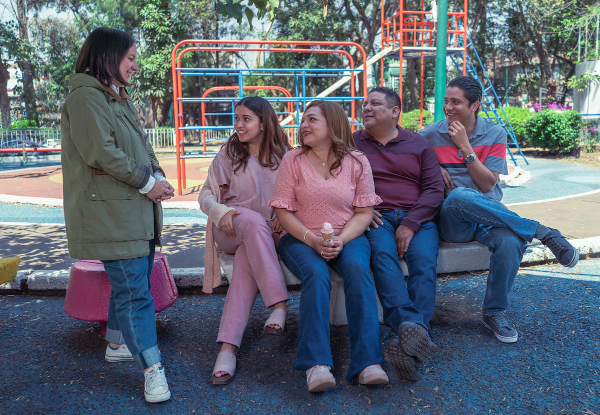 Five adults socializing in a playground, sitting and standing by colorful playground equipment, surrounded by trees and greenery on a sunny day.