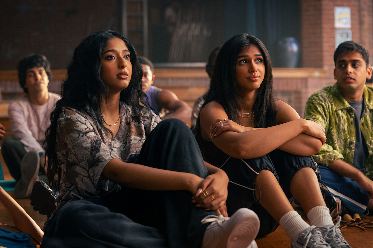 A group of teenagers sitting on a gym floor, attentively listening, with two girls in the foreground and other students in the background in a school setting.