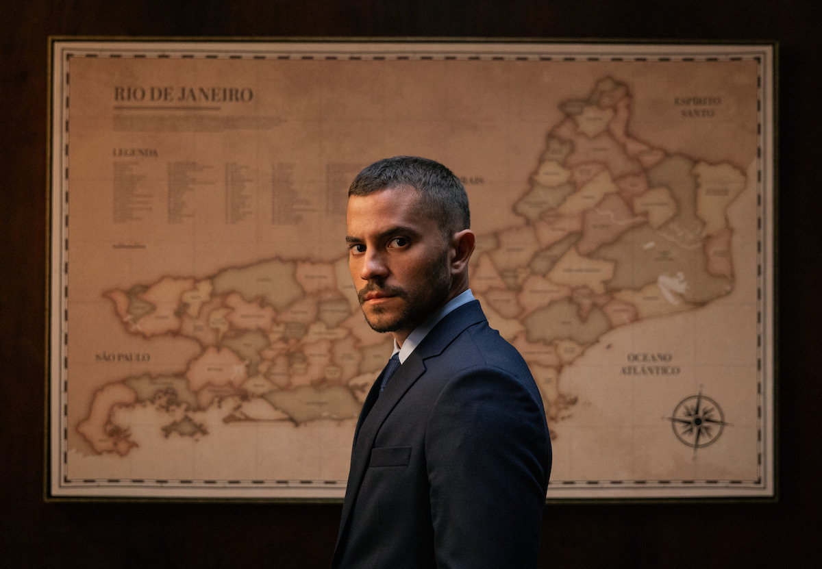 Ator André Lamoglia posa de terno em frente a um mapa do Rio de Janeiro, em cena da série “Donos do Jogo”
