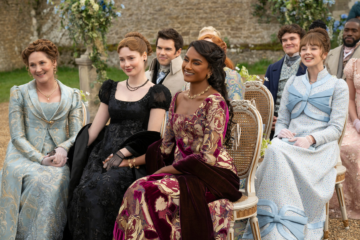 A group of people in elegant period costumes sit on chairs outdoors at what appears to be a formal event or ceremony, surrounded by flowers and stone walls in a garden setting.
