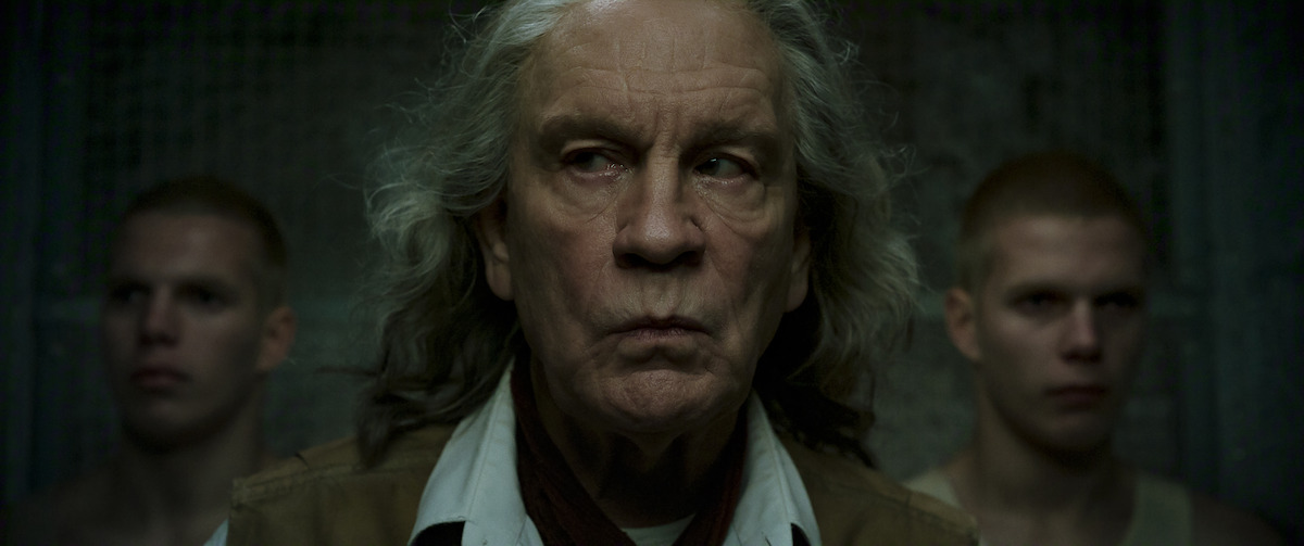 Older man with long gray hair stands in the center, looking serious, flanked by two young men with shaved heads. The setting is dimly lit, with a gritty, industrial or prison-like background.
