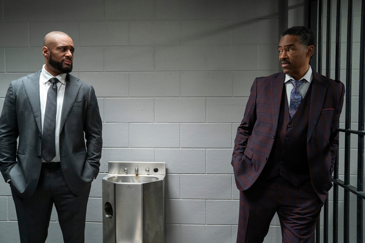 Two men in suits stand in a jail or prison environment, facing each other with serious expressions. One stands by a water fountain, and the other is near metal bars, suggesting a tense conversation in a holding area or cell block.