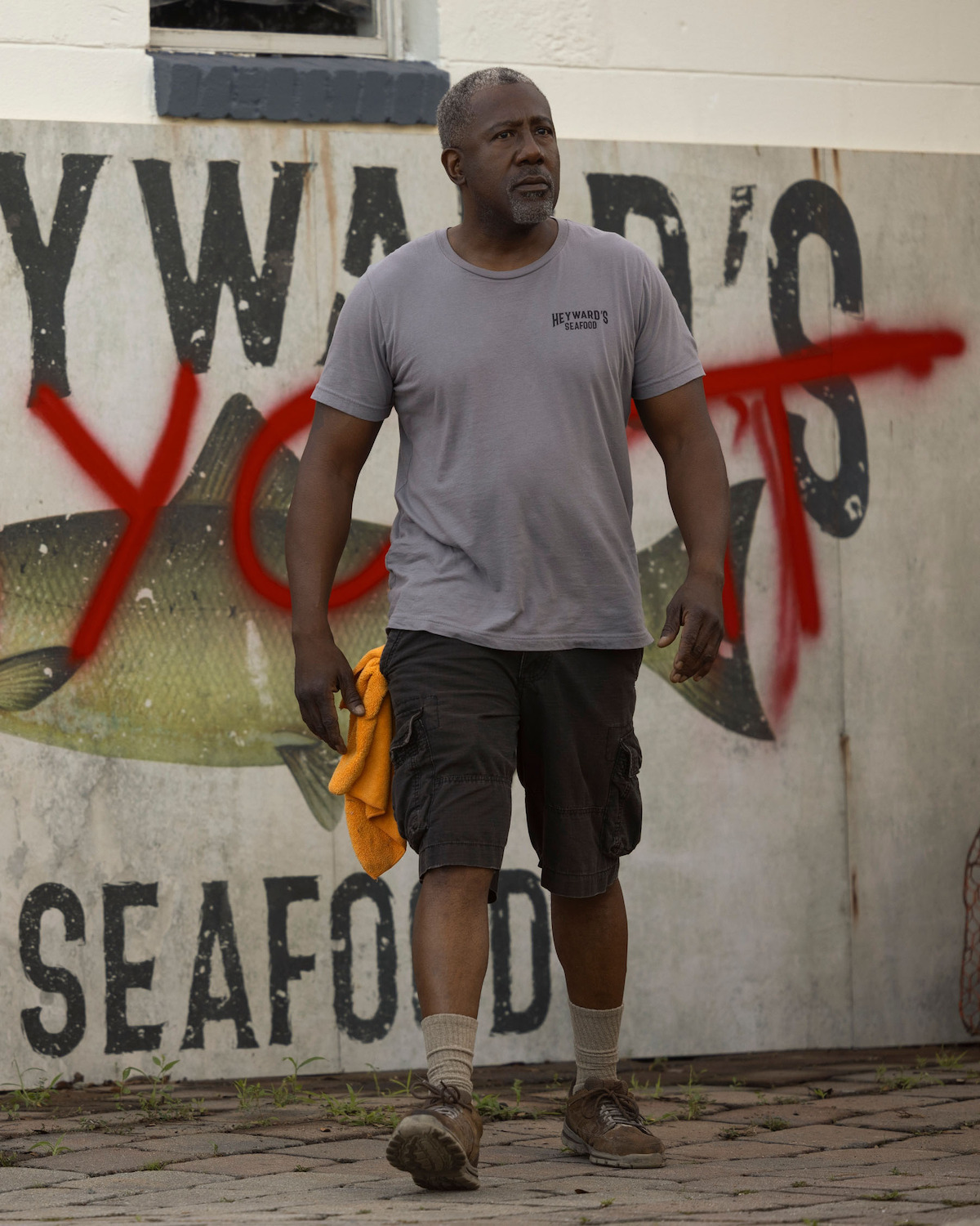 E. Roger Mitchell as Bobby Heyward, in casual clothes holding a yellow cloth walks on cobblestone in front of a seafood restaurant mural with graffiti, suggesting an urban or small town setting.