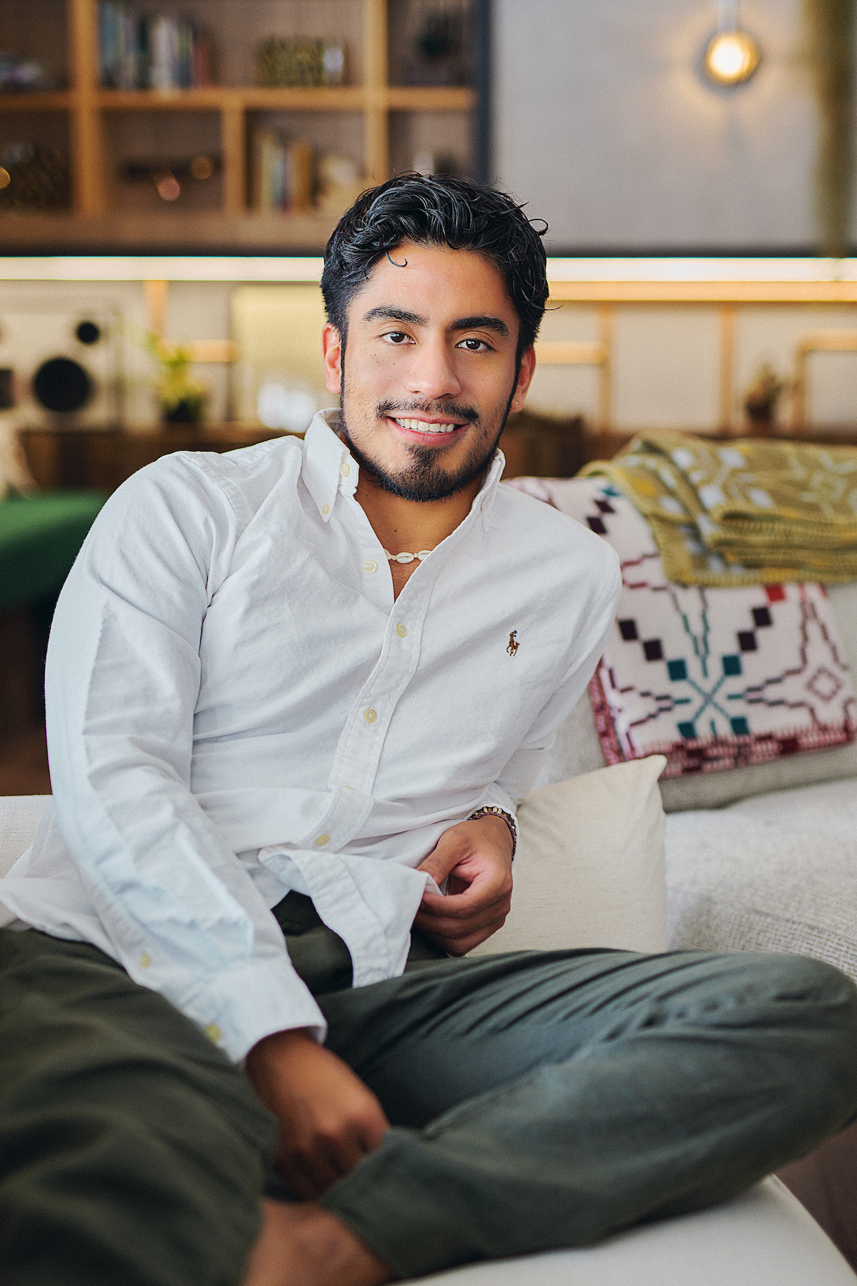 A young man in a white shirt sits relaxed on a modern sofa in a cozy, well-lit living room with shelves, decor, and patterned blankets, creating a warm and inviting mood.