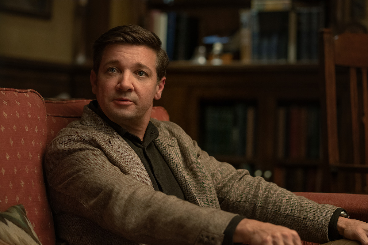 Man in a brown blazer sits on a red couch in a cozy, warmly lit library or study, surrounded by bookshelves, creating an atmosphere of calm conversation or thoughtful reflection.