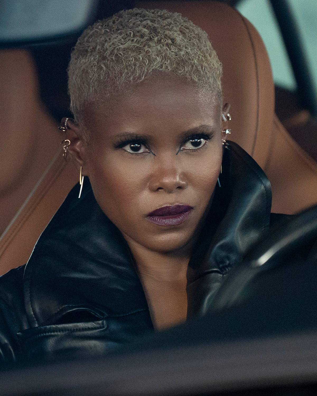 Woman with short blonde hair wearing a black leather jacket, sitting in a brown leather car interior, looking forward with a serious expression.