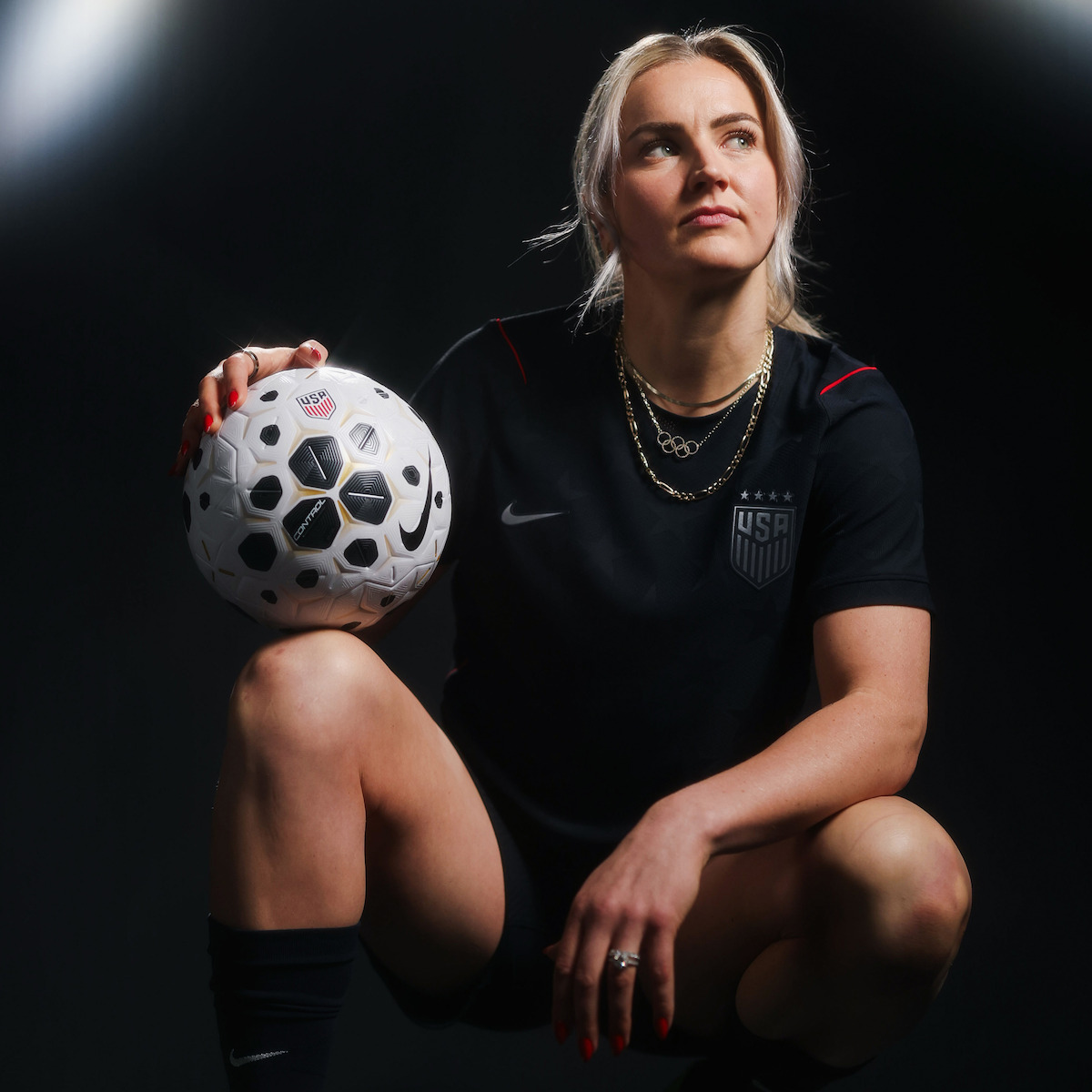 Female soccer player in a black USA uniform poses confidently in a dark studio, kneeling with one hand on her knee and the other resting on a soccer ball, dramatic lighting highlighting her determined expression.
