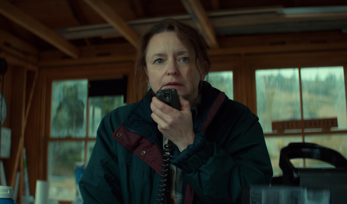 Woman in a heavy jacket holding a radio, inside a wooden cabin with large windows showing an outdoor rural landscape, looks concerned or focused while communicating.