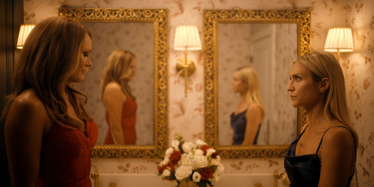 Two women in elegant dresses stand facing each other in a warmly lit, ornate bathroom with floral wallpaper, gold-framed mirrors, wall lamps, and a vase of flowers on the counter, their reflections visible in the mirrors.