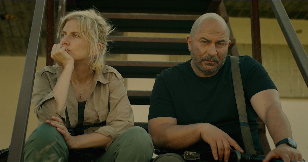 Two serious people sit on outdoor metal stairs in casual, tactical clothing, appearing tired or contemplative, with sunlight casting shadows behind them against a plain wall in a rugged environment.