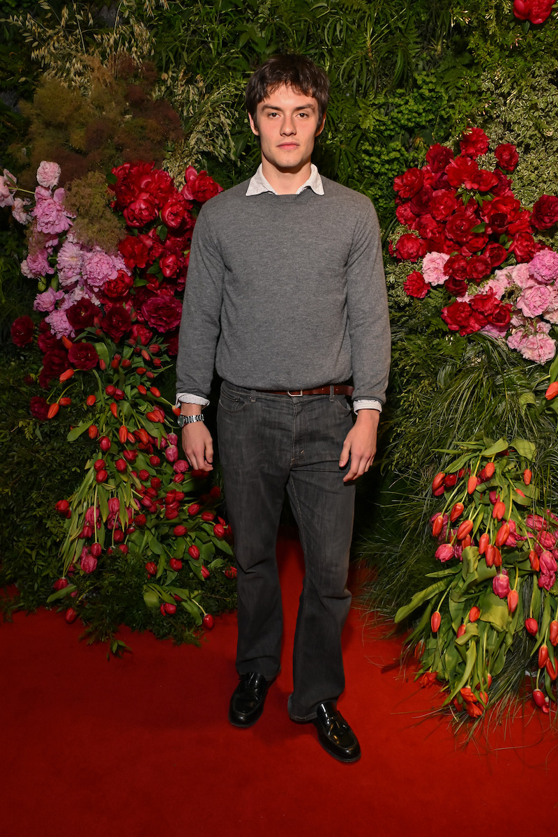 A man in a gray sweater and dark jeans stands on a red carpet surrounded by lush greenery and vibrant red and pink flowers arranged as a backdrop.