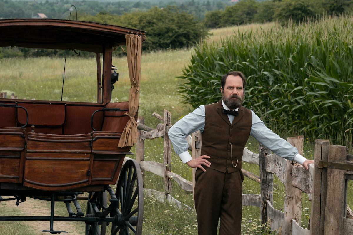 A man in vintage clothing stands by a wooden fence and carriage in a rural setting with a field and corn crops, evoking a calm, historical atmosphere.