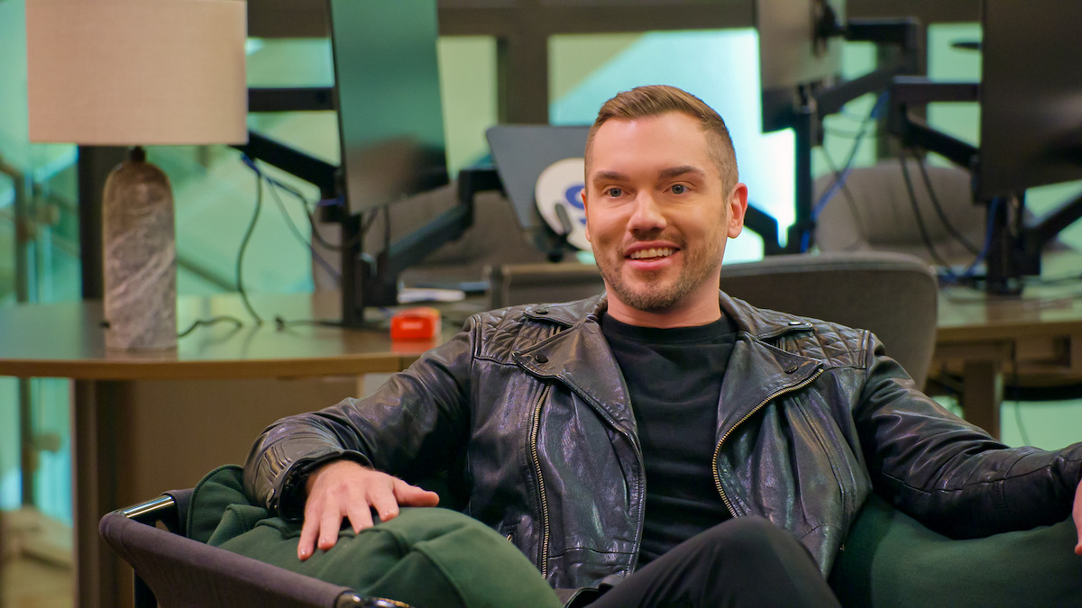 Man in black leather jacket sits relaxed on a green couch in a modern office, with desk, lamp, and computer screens in the background, creating a casual and upbeat mood.