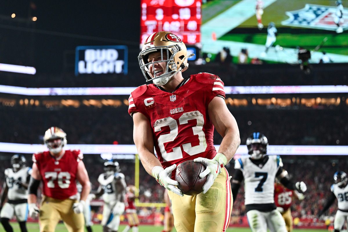 San Francisco 49ers player celebrating a touchdown in a football stadium during a night game, holding the ball with teammates and opposing players in the background, under bright stadium lights.