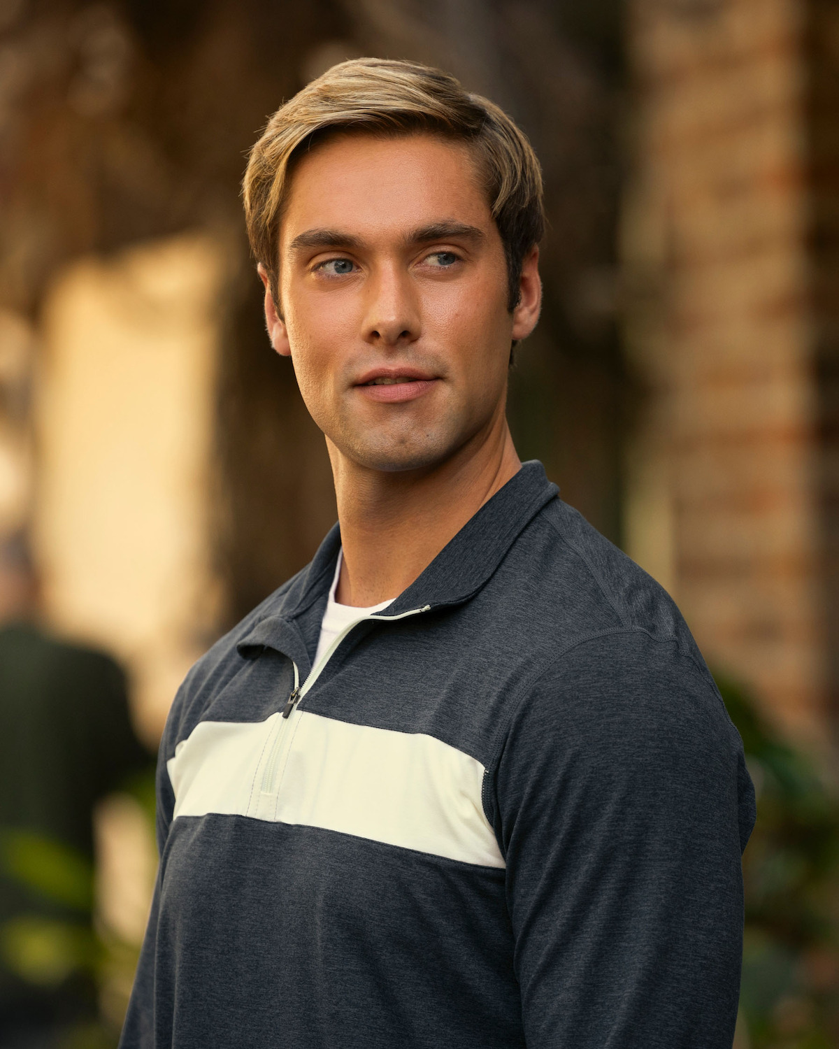 Austin North as Topper, with light hair wearing a gray zip-up shirt stands outdoors near a brick wall, looking to the side with a relaxed expression. The background is softly blurred, hinting at plants and another person.