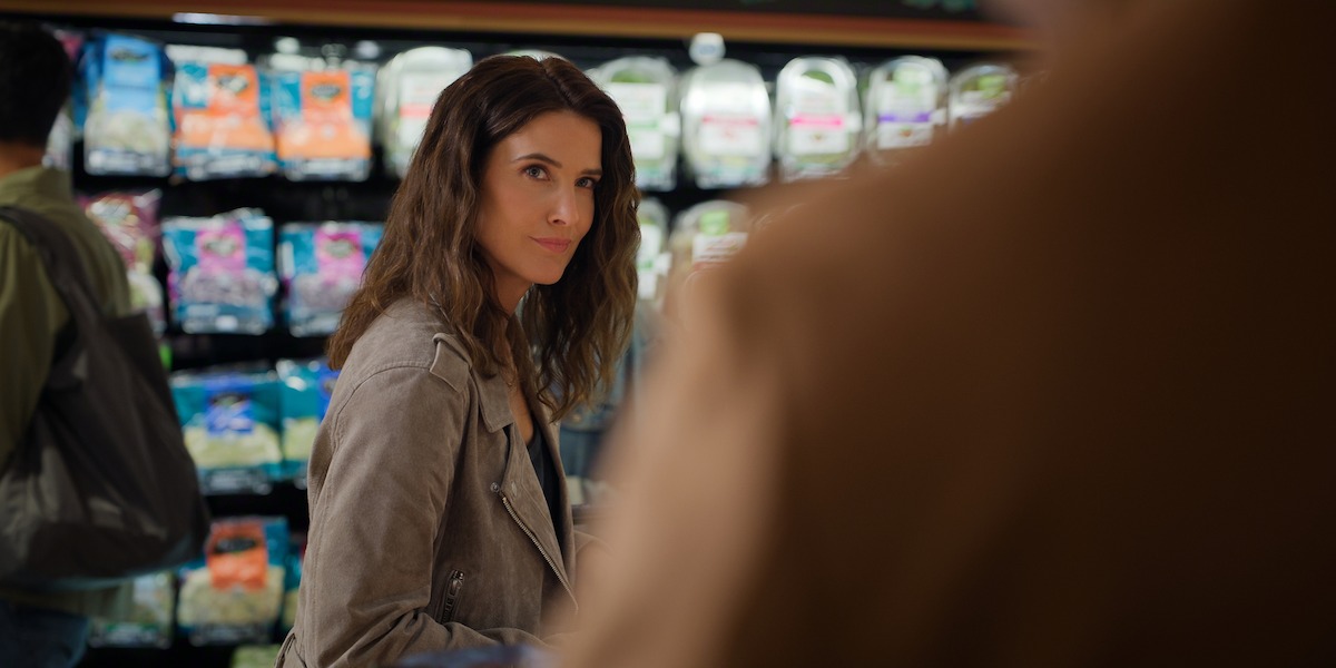 Woman with long brown hair wearing a light jacket stands in a grocery store produce aisle, looking at a person in the foreground, with bags of salad on shelves behind her.