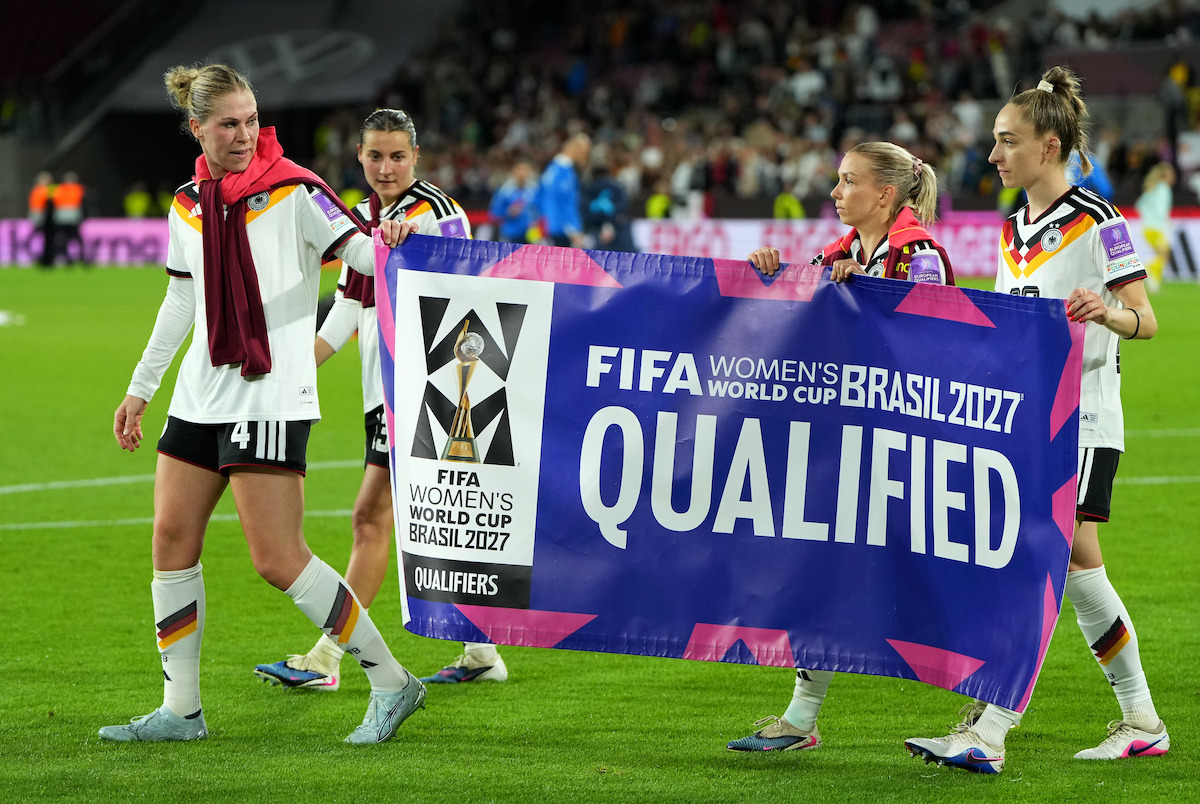 Female soccer players on a field celebrate qualification for the FIFA Women’s World Cup 2027 in Brazil, holding a large banner stating "Qualified" with a stadium and crowd in the background.