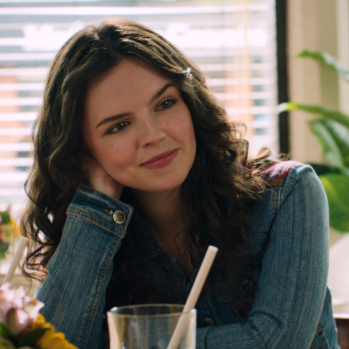 A young woman with curly brown hair in a denim jacket sits at a table in a sunlit cafe, resting her head on her hand and smiling, with a drink and flowers in front of her, window blinds in the background.