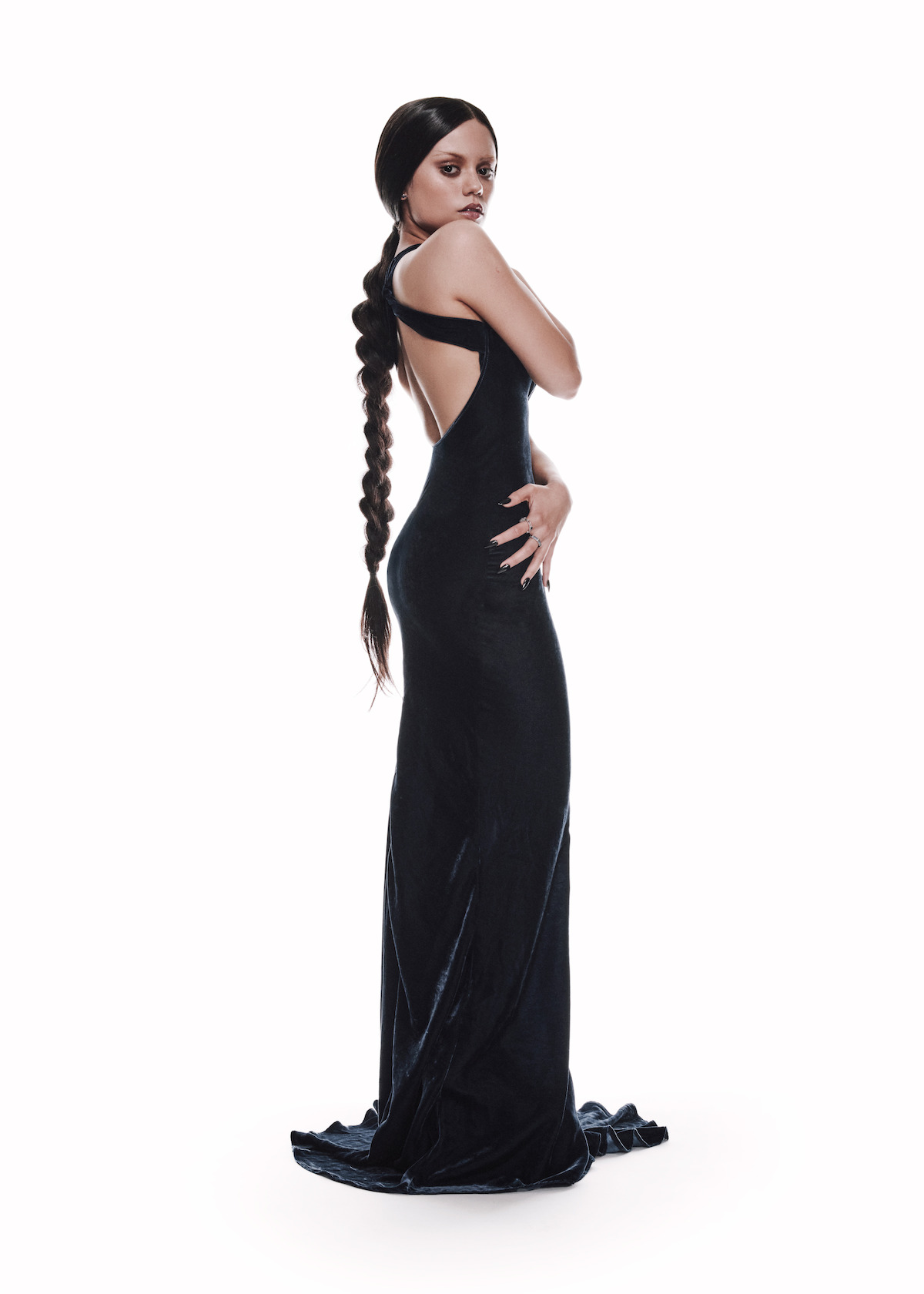 Jenna Ortega stands on a bright white background looking back over her shoulder wearing a black backless dress that gathers on the floor around her feet, hair in a long braid extending below her back.